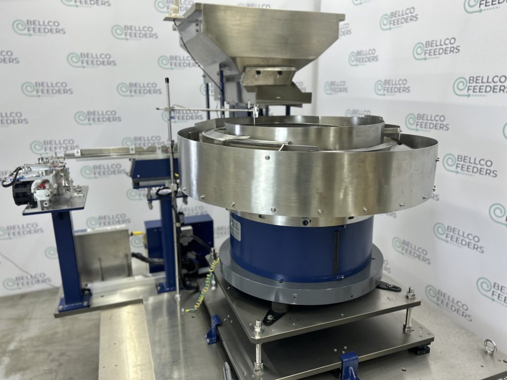 1100mm stainless steel Bellco vibratory bowl with a conveyor-fed hopper above it, built to feed machined metal components