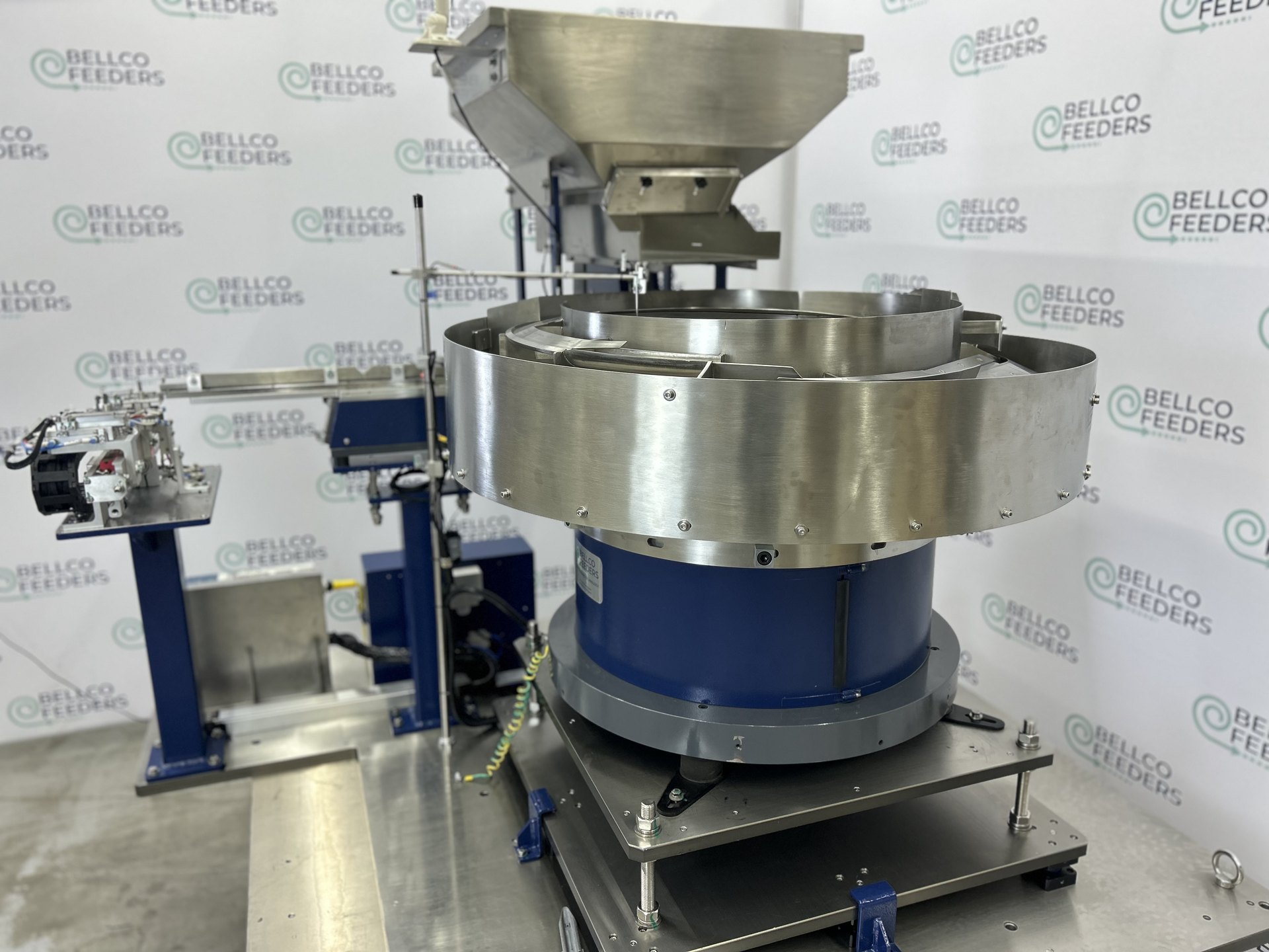 1100mm stainless steel Bellco vibratory bowl with a conveyor-fed hopper above it, built to feed machined metal components