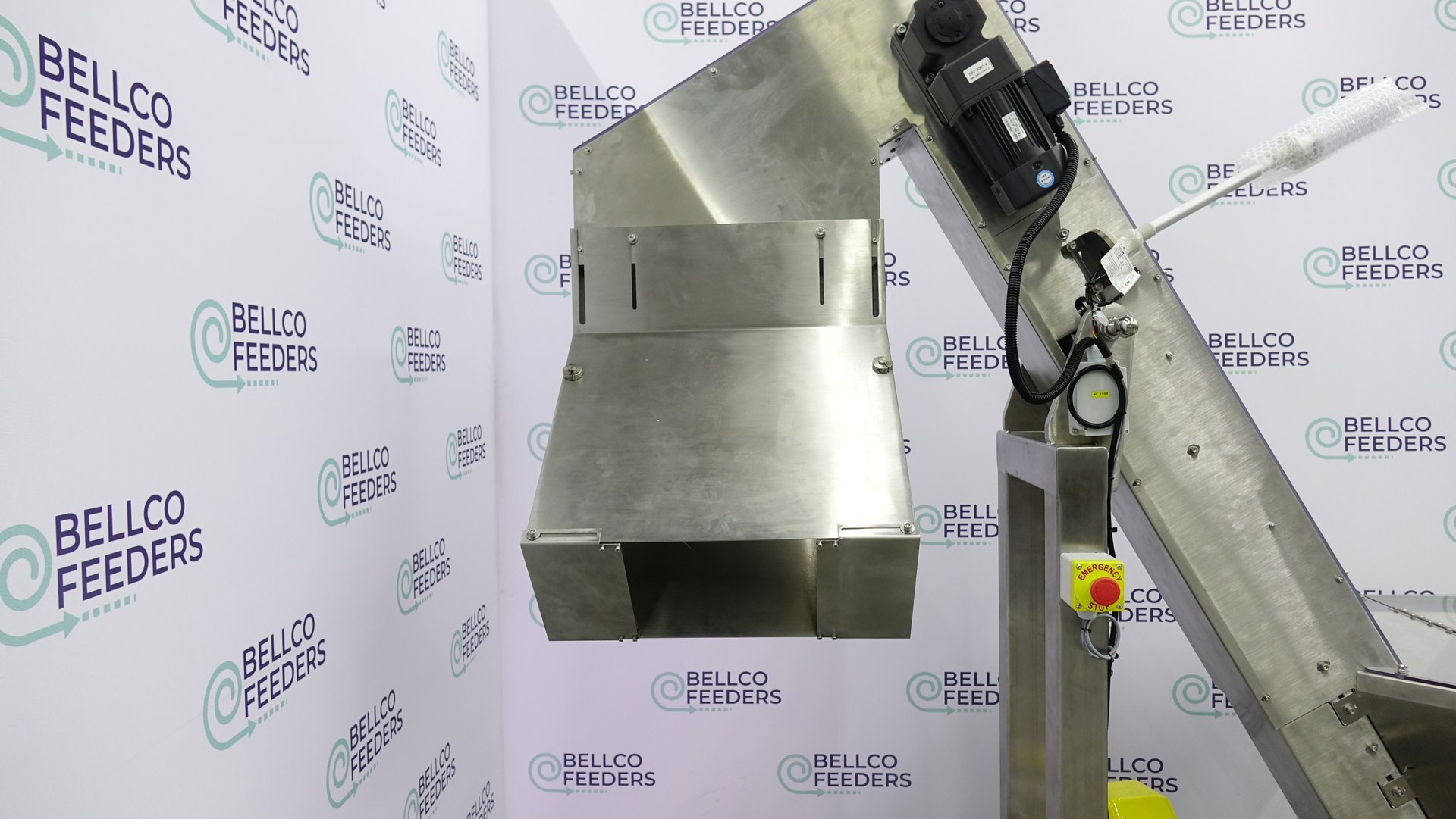 Discharge chute on a Bellco elevated conveyor hopper, angled to drop parts into a feeder bowl