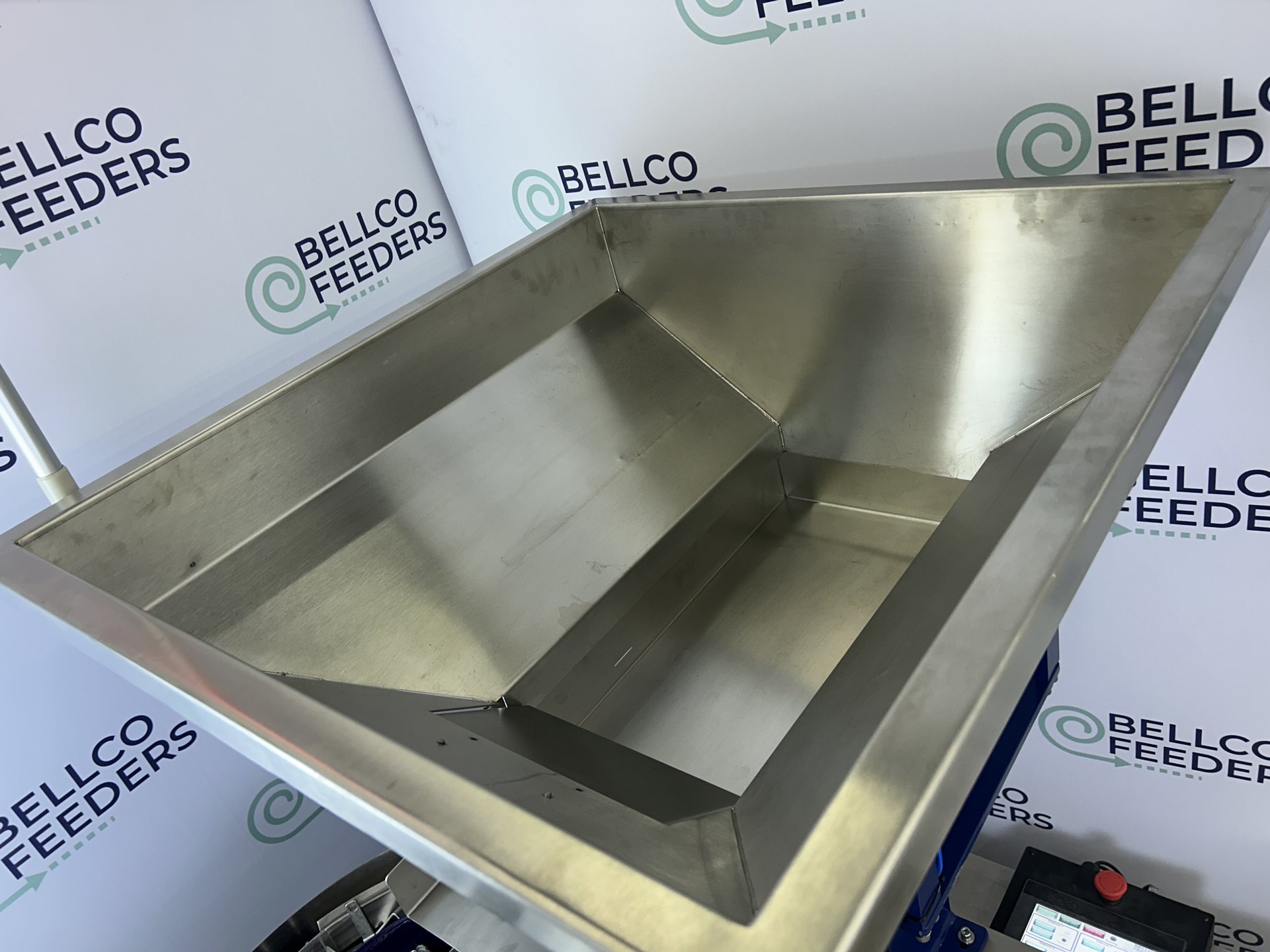 Empty stainless vibratory hopper built by Bellco, shown against the Bellco backdrop