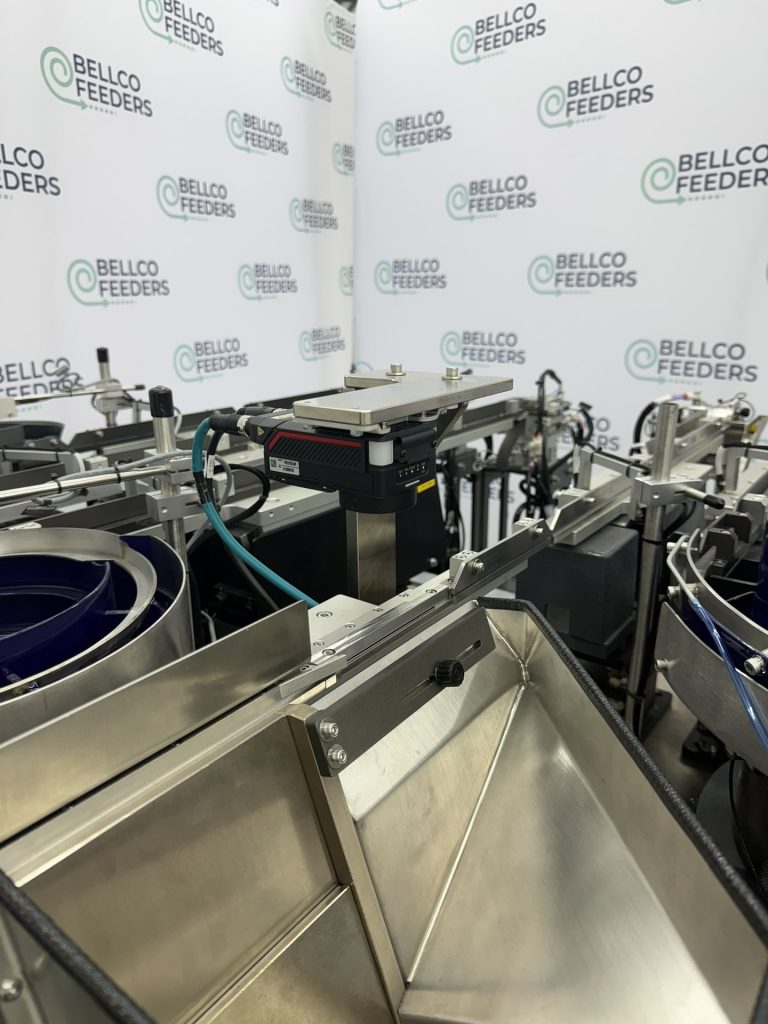 Tabletop Bellco step feeder with a stainless loading hopper, built to sit inside an existing robotic assembly cell