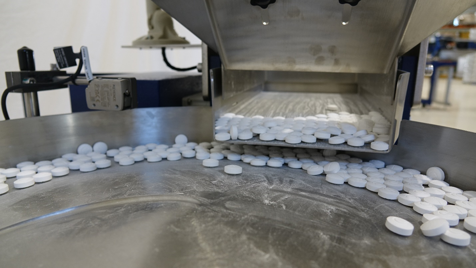 White tablets traveling along the track of a stainless feeder bowl below the hopper discharge