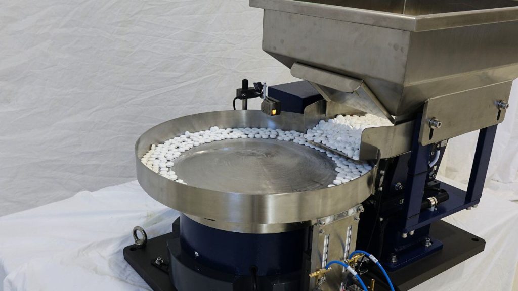 Polished stainless vibratory bowl feeder with an overhead hopper, feeding white tablets