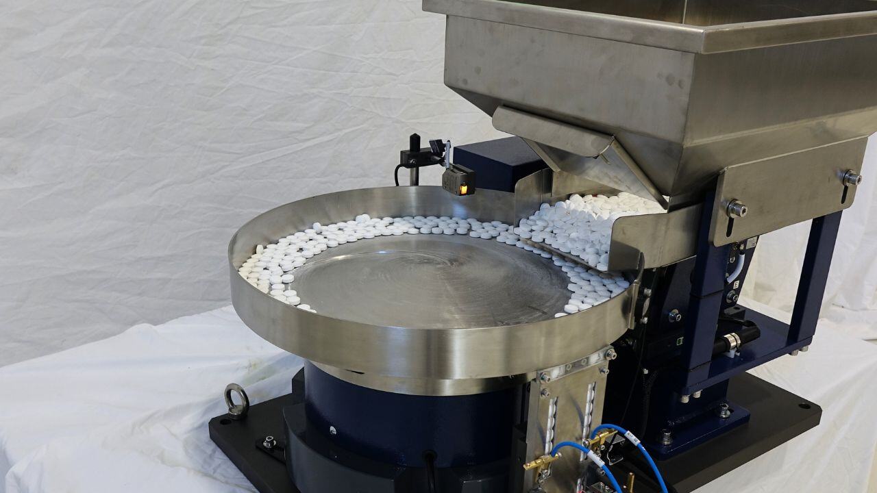 Polished stainless vibratory bowl feeder with an overhead hopper, feeding white tablets